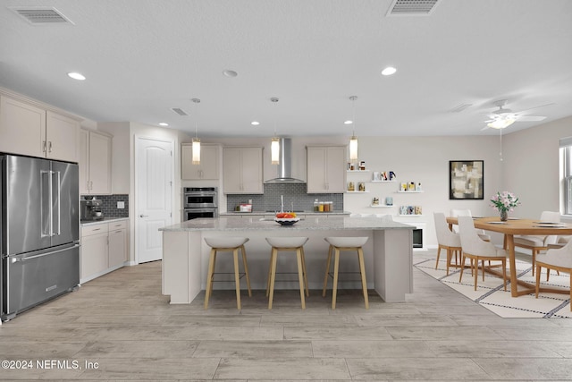 kitchen featuring a center island with sink, wall chimney exhaust hood, light stone counters, and appliances with stainless steel finishes