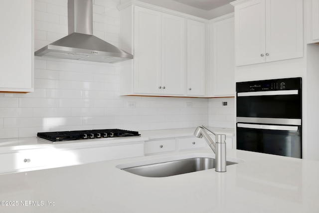 kitchen featuring sink, white cabinets, backsplash, black double oven, and wall chimney range hood