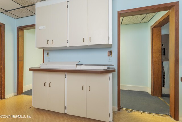 kitchen featuring a drop ceiling, white cabinets, light carpet, and electric panel