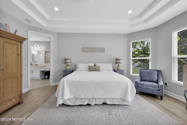 bedroom with a raised ceiling, light wood-type flooring, and ensuite bath