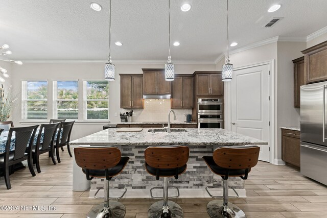 kitchen featuring hanging light fixtures, light stone counters, stainless steel appliances, sink, and a center island with sink