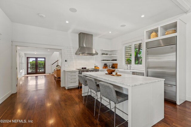 kitchen featuring built in fridge, a kitchen island, a breakfast bar, dark wood-type flooring, and wall chimney range hood