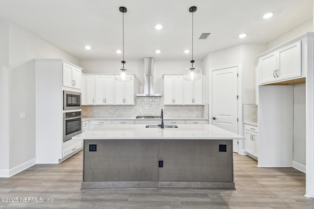 kitchen featuring wall chimney range hood, a kitchen island with sink, black microwave, white cabinets, and stainless steel oven