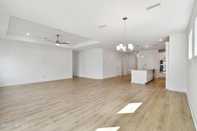 unfurnished living room featuring ceiling fan with notable chandelier, sink, a raised ceiling, and light hardwood / wood-style floors