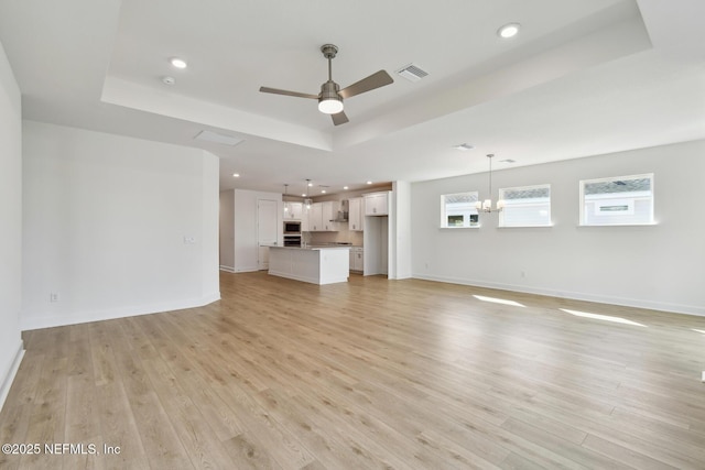 unfurnished living room featuring a raised ceiling, ceiling fan with notable chandelier, and light hardwood / wood-style flooring