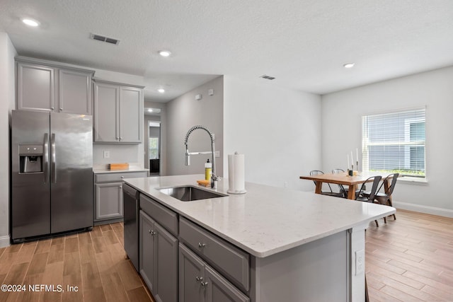 kitchen with stainless steel appliances, light hardwood / wood-style floors, light stone counters, a kitchen island with sink, and gray cabinets