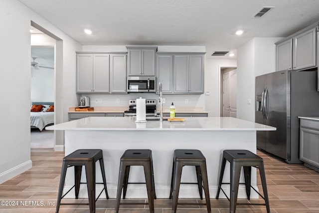 kitchen with light wood-type flooring, an island with sink, gray cabinets, and stainless steel appliances