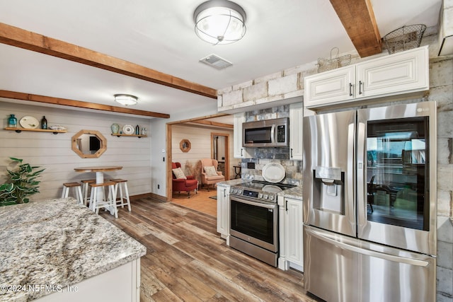 kitchen with appliances with stainless steel finishes, beamed ceiling, white cabinetry, hardwood / wood-style flooring, and light stone countertops