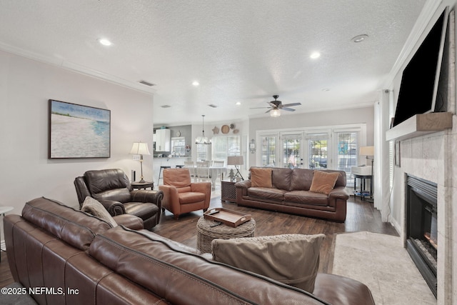 living room with ceiling fan with notable chandelier, crown molding, a textured ceiling, wood-type flooring, and a tiled fireplace