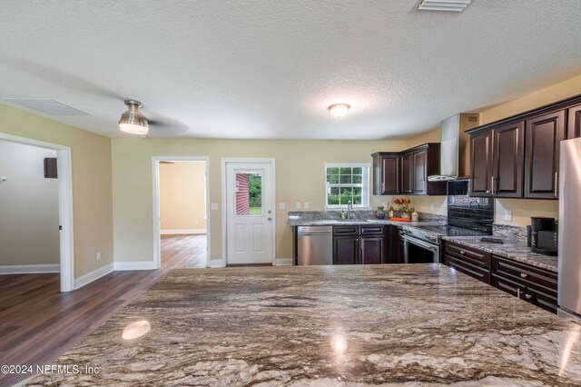 kitchen featuring wall chimney range hood, dark hardwood / wood-style flooring, stone countertops, stainless steel appliances, and dark brown cabinets