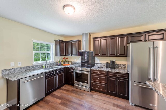 kitchen featuring stainless steel appliances, sink, light stone countertops, wall chimney range hood, and hardwood / wood-style flooring