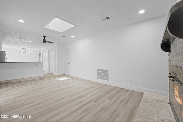 unfurnished living room with a fireplace, a textured ceiling, a skylight, ceiling fan, and light hardwood / wood-style flooring