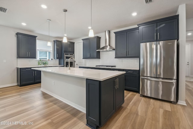 kitchen featuring stainless steel appliances, a kitchen island with sink, sink, wall chimney range hood, and pendant lighting