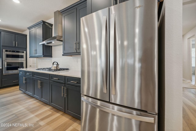 kitchen featuring light wood finished floors, stainless steel appliances, tasteful backsplash, light countertops, and wall chimney range hood