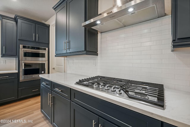 kitchen with light stone counters, light wood-style flooring, exhaust hood, appliances with stainless steel finishes, and tasteful backsplash