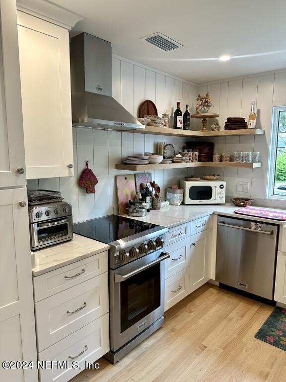 kitchen with wall chimney range hood, light wood-type flooring, white cabinetry, appliances with stainless steel finishes, and light stone counters