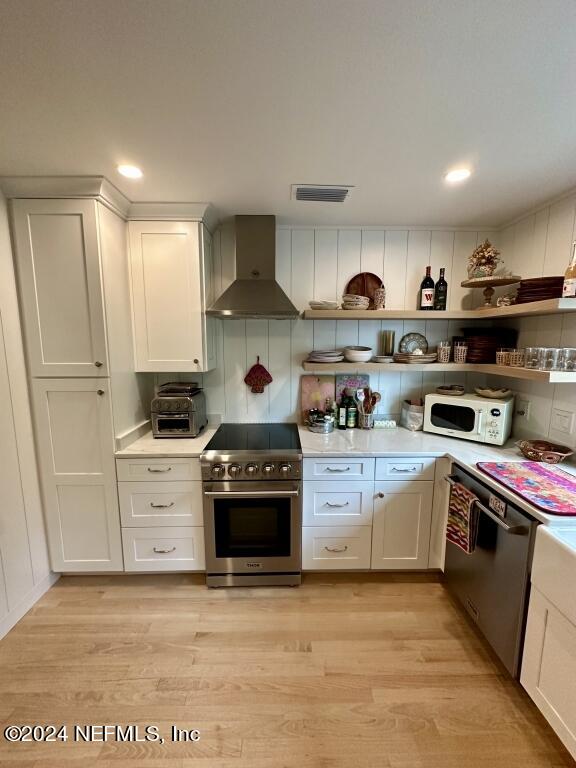 kitchen with wall chimney range hood, white cabinets, light hardwood / wood-style floors, and stainless steel appliances