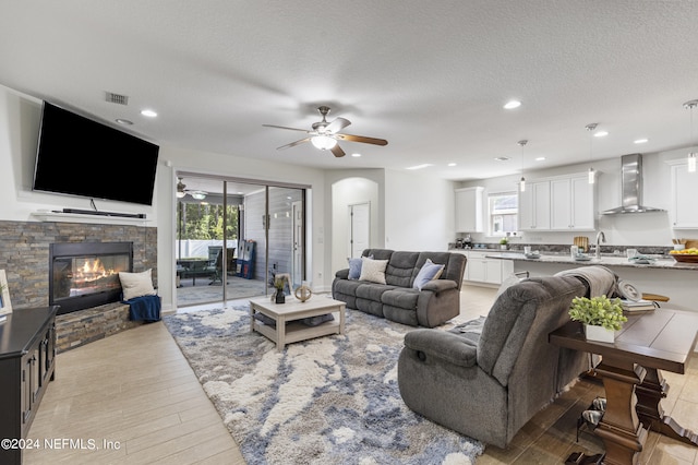 living area featuring visible vents, a ceiling fan, a textured ceiling, a stone fireplace, and light wood-type flooring