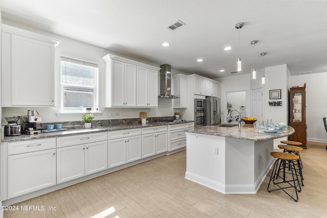 kitchen featuring visible vents, an island with sink, wall chimney range hood, white cabinetry, and a sink