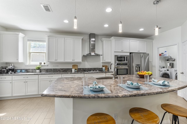 kitchen featuring stainless steel appliances, wall chimney range hood, a center island with sink, and decorative light fixtures