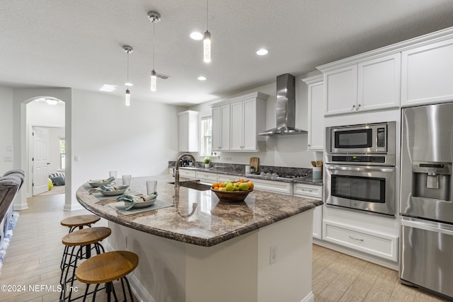 kitchen with a center island with sink, arched walkways, wall chimney exhaust hood, hanging light fixtures, and stainless steel appliances