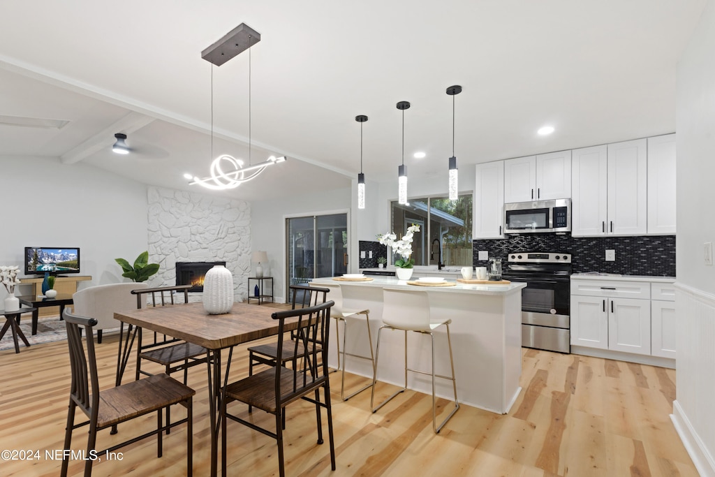 dining area featuring vaulted ceiling with beams, light wood-type flooring, a stone fireplace, ceiling fan with notable chandelier, and sink