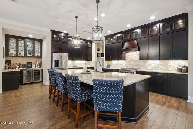 kitchen with crown molding, appliances with stainless steel finishes, custom range hood, a center island with sink, and dark brown cabinetry