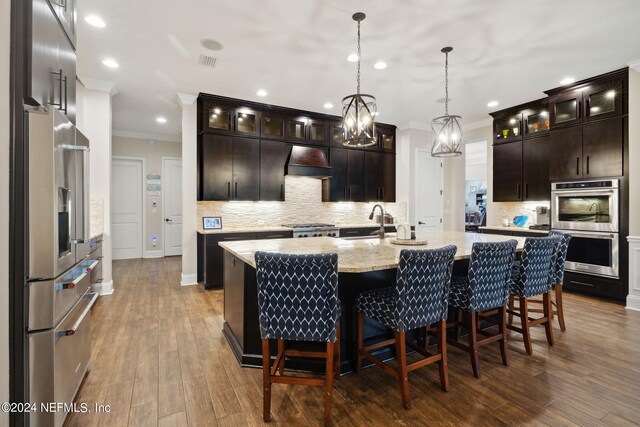 kitchen with stainless steel appliances, an island with sink, light stone countertops, dark wood-type flooring, and custom range hood