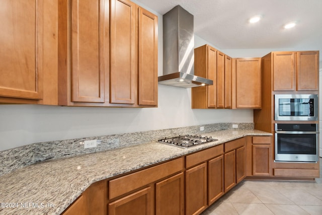 kitchen featuring light stone counters, light tile patterned flooring, appliances with stainless steel finishes, wall chimney range hood, and brown cabinetry