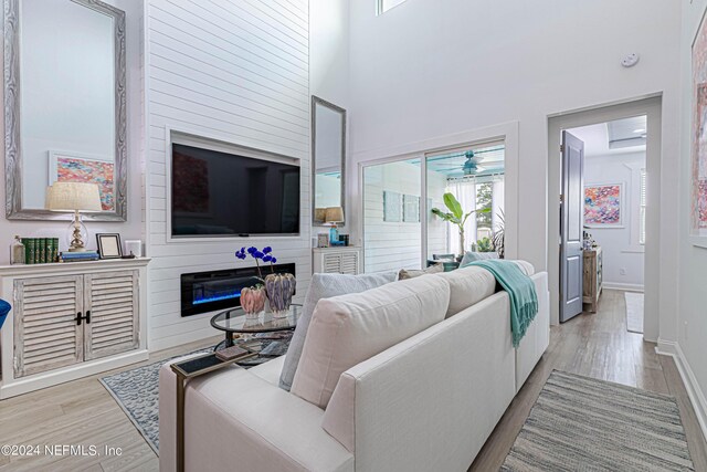living area with baseboards, ceiling fan, a towering ceiling, light wood-style floors, and a glass covered fireplace