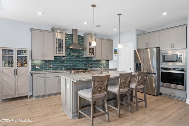 kitchen featuring a kitchen island with sink, stainless steel appliances, wall chimney range hood, and light hardwood / wood-style floors