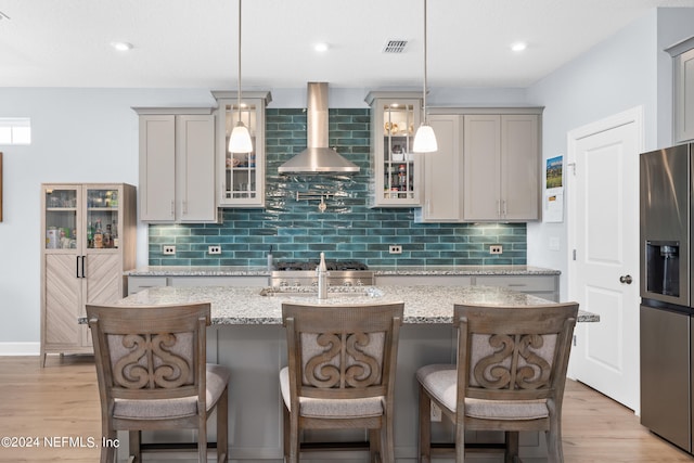 kitchen featuring hanging light fixtures, light stone counters, wall chimney range hood, an island with sink, and stainless steel refrigerator with ice dispenser