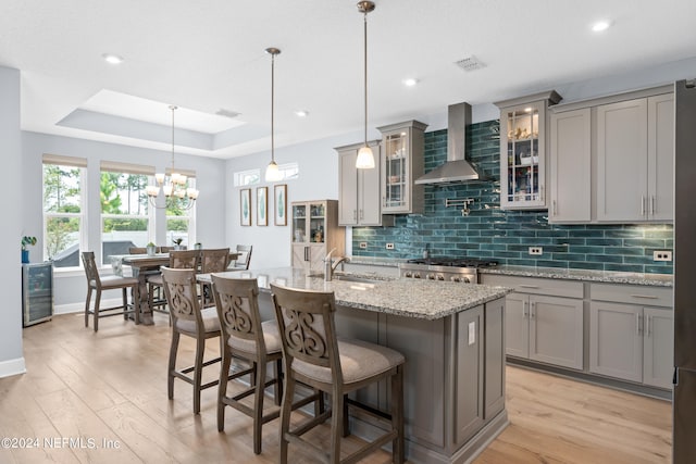 kitchen featuring light wood-type flooring, an inviting chandelier, hanging light fixtures, wall chimney exhaust hood, and a center island with sink