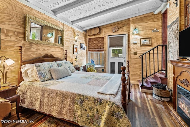 bedroom with beam ceiling, a glass covered fireplace, wood walls, and wood finished floors