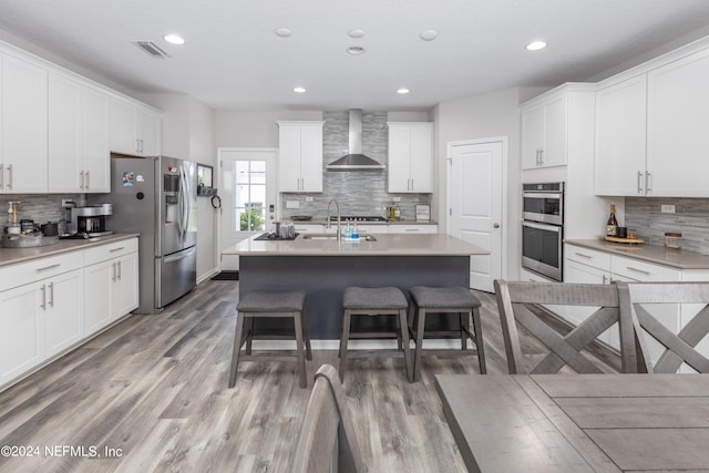 kitchen with a kitchen island with sink, white cabinetry, stainless steel appliances, wall chimney exhaust hood, and hardwood / wood-style flooring