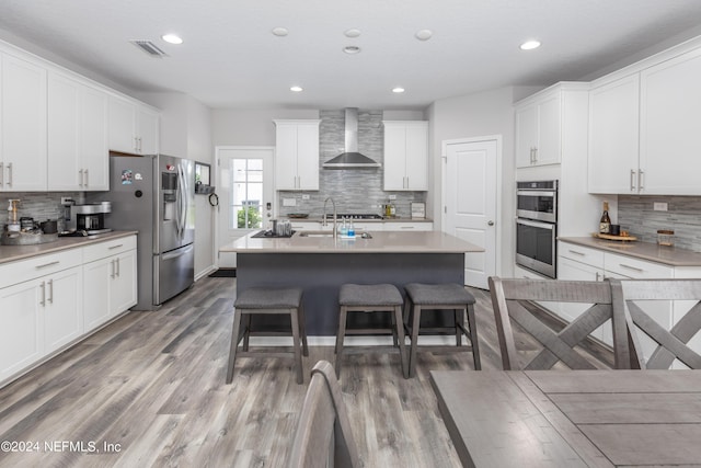 kitchen featuring wall chimney range hood, light wood-style flooring, a kitchen island with sink, appliances with stainless steel finishes, and a kitchen breakfast bar