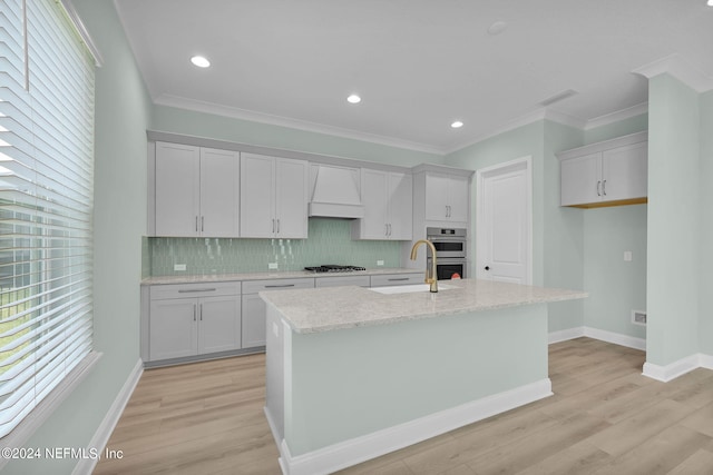 kitchen with a kitchen island with sink, stainless steel double oven, premium range hood, gas cooktop, and a sink