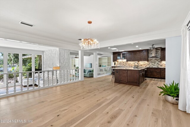 interior space with an inviting chandelier, a center island with sink, backsplash, light hardwood / wood-style floors, and wall chimney range hood