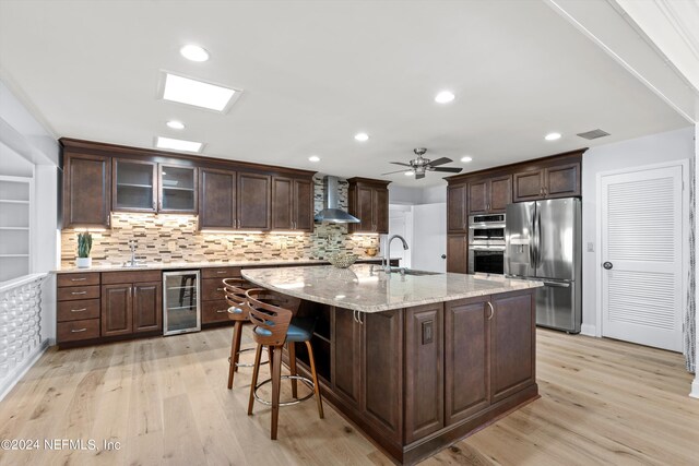 kitchen featuring a kitchen island with sink, beverage cooler, wall chimney exhaust hood, appliances with stainless steel finishes, and light hardwood / wood-style floors