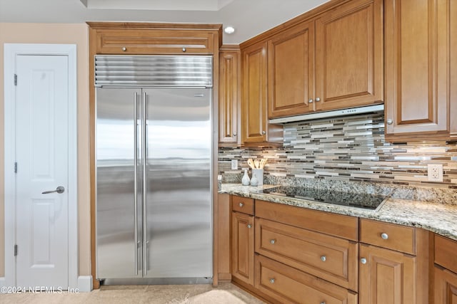 kitchen featuring built in refrigerator, light stone countertops, black electric cooktop, under cabinet range hood, and backsplash