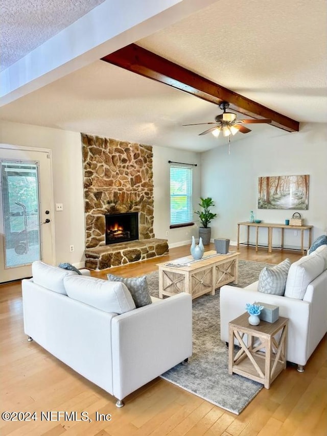 living room with a textured ceiling, lofted ceiling with beams, a stone fireplace, a ceiling fan, and light wood-type flooring