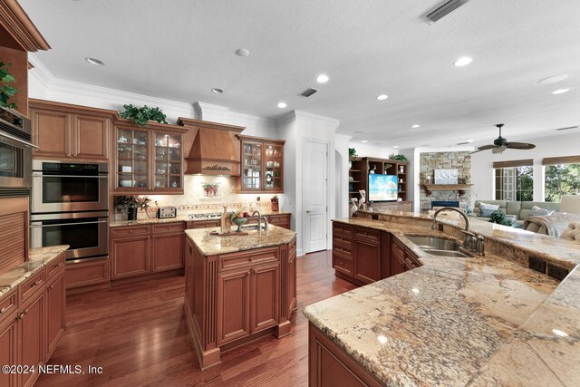 kitchen with a fireplace, custom exhaust hood, sink, double oven, and a kitchen island with sink