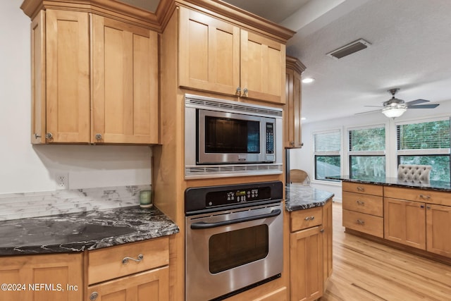 kitchen with appliances with stainless steel finishes, dark stone countertops, ceiling fan, and light hardwood / wood-style flooring