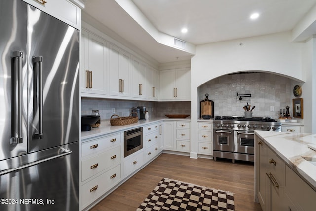 kitchen with built in appliances, wood-type flooring, tasteful backsplash, white cabinetry, and light stone counters