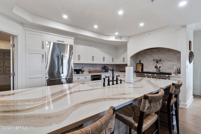 kitchen with white cabinets, stainless steel built in fridge, light wood-type flooring, light stone counters, and decorative backsplash