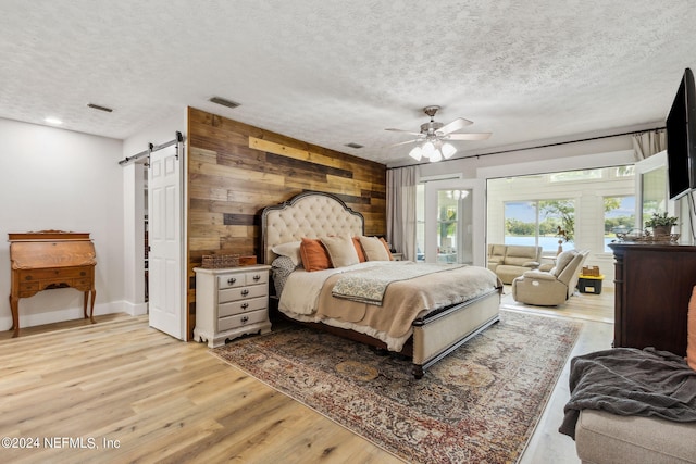 bedroom featuring a textured ceiling, light hardwood / wood-style flooring, wood walls, ceiling fan, and a barn door