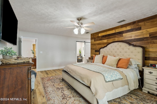 bedroom featuring a textured ceiling, light hardwood / wood-style flooring, a barn door, ceiling fan, and wooden walls