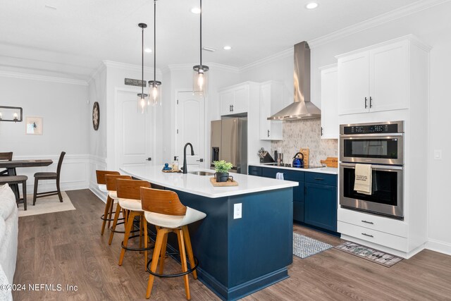 kitchen with decorative light fixtures, stainless steel appliances, an island with sink, dark wood-type flooring, and wall chimney exhaust hood