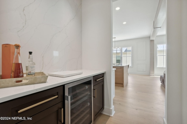 bar with wine cooler, light stone countertops, light hardwood / wood-style floors, dark brown cabinetry, and decorative light fixtures