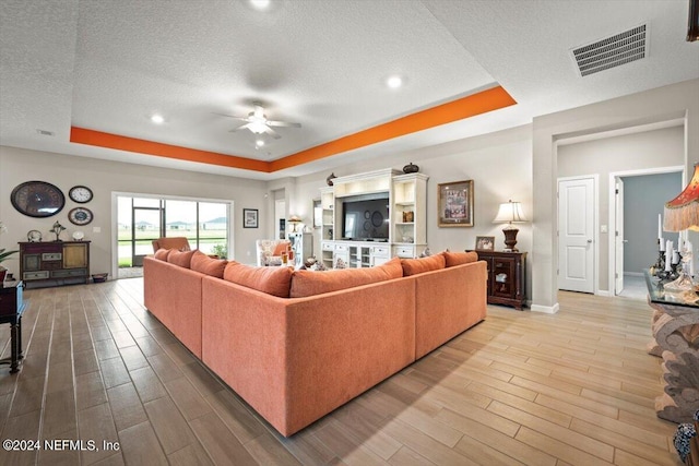 living room with a textured ceiling, ceiling fan, a tray ceiling, and light hardwood / wood-style floors